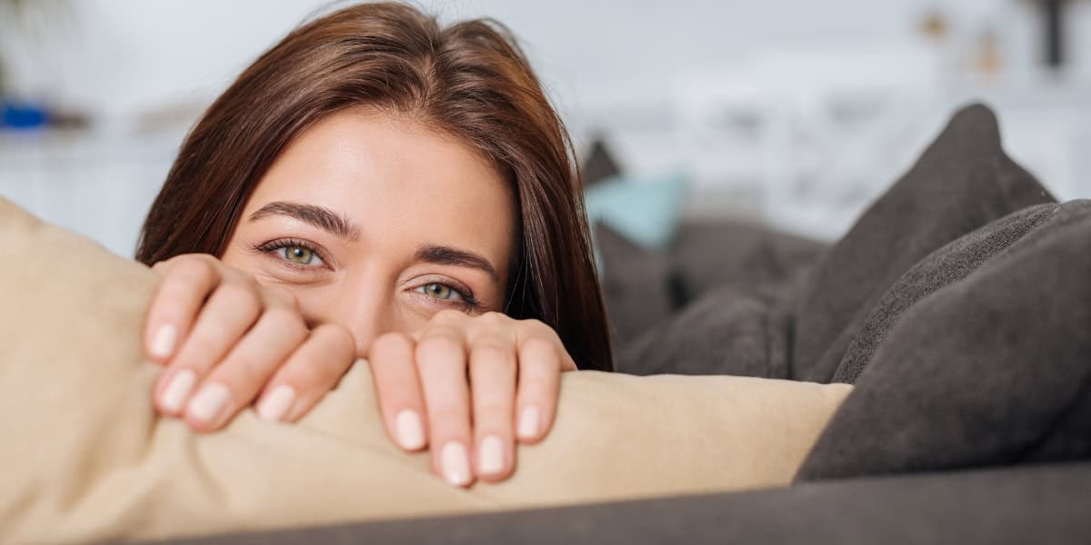 A woman is hiding behind a pillow on a couch.