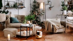 Cozy living room with beige furniture, green plants, books, and candles on a round table. Neutral tones and natural lighting create a warm atmosphere.