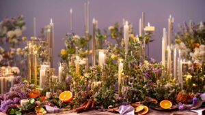 A table adorned with tall candles, lush greenery, clusters of purple flowers, sliced oranges, and cinnamon sticks.