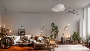 A spacious living room with a beige sectional sofa, plants, candles, and soft lighting. A modern cloud shaped light fixture is on the ceiling. Cozy and minimalist decor.