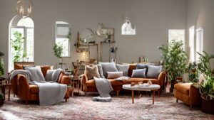 A cozy living room with brown sofas, gray blankets, a round coffee table, various potted plants, and multiple wall mirrors. Natural light streams in through arched windows.