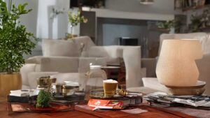 Cozy living room with a wooden table holding a lamp, books, and coffee cup. Cream colored sofas and greenery in the background.