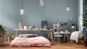 Cozy bedroom with a low bed, desk, and chair. Soft pink bedding, green plants, and minimalist decor against a muted blue wall.