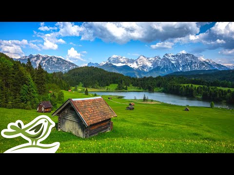 A small wooden cabin sits on a grassy field near a lake, with scattered huts, dense forest, and snow capped mountains in the background under a partly cloudy sky.