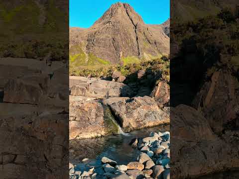 Rocky waterfall flows into a small pool, surrounded by large stones, with a steep, rugged mountain rising in the background under a clear blue sky.