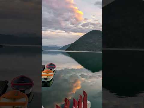 Still lake with reflections of mountains and a cloudy sky at sunset; small boats are docked in the foreground near a red fence.